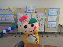 掲示物の前で両頬に手を添えたポーズを決めているかまたんの写真